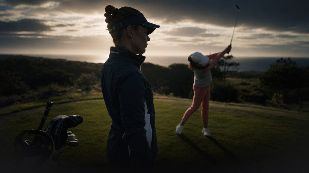 Two golfers on a coastal golf course at sunset, one watching while the other swings a club in the background with a cart nearby.